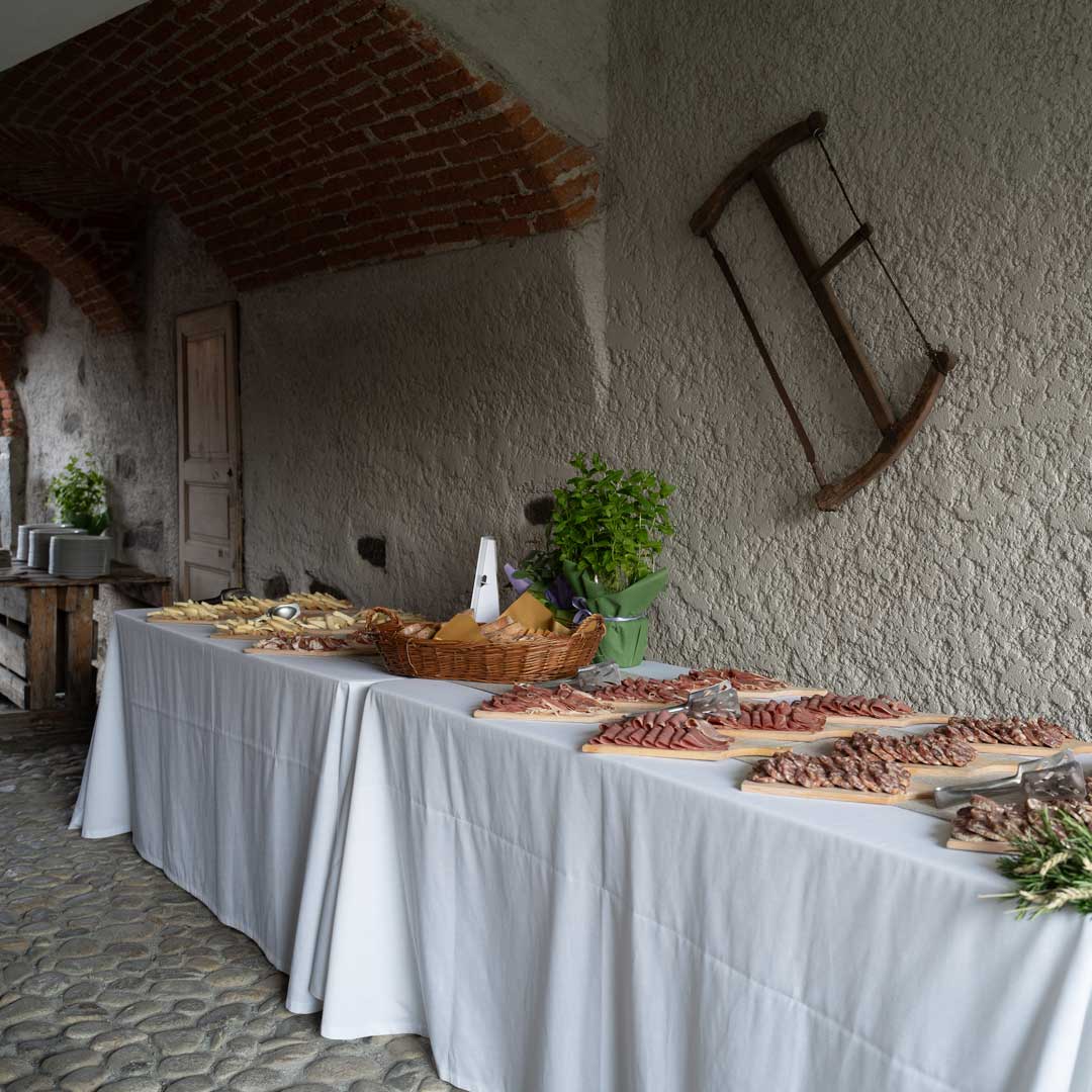 L'immagine mostra un ricco buffet allestito all'aperto, in un contesto rustico e suggestivo. Il tavolo, coperto da una tovaglia bianca, è imbandito con una varietà di salumi affettati, formaggi e pane, disposti su taglieri di legno e cesti di vimini. Sullo sfondo, si notano pareti in pietra, con un arco in mattoni a vista e un'antica sega appesa al muro. Un vaso con una pianta verde e fiori colorati completa l'allestimento.
Assapora i sapori autentici della tradizione con il servizio di catering di Just Grilled Catering by Aglietti. Scopri i nostri menu personalizzati e richiedere un preventivo.