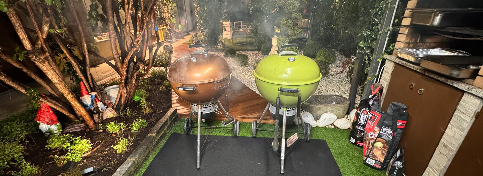 un'immagine di un'area esterna pronta per un evento di Hust Grilled Catering by AGlietti. Due barbecue a carbone di tipo kettle, uno marrone ramato e l'altro verde lime, sono posizionati su un tappeto nero e un prato sintetico al centro della scena. Il cortile è caratterizzato da ghiaia chiara, una siepe di arbusti verdi con nani da giardino in ceramica, vetrate verticali, sedute basse con cuscini chiari in mezzo ad un gazebo ricoperto di piante, un breve passaggio pedonale in legno e confezioni di carbone e utensili pronti all'uso. Organizza il tuo evento perfetto con Just Grilled Catering by Aglietti! Scopri i nostri menu personalizzati e richiedere un preventivo gratuito.