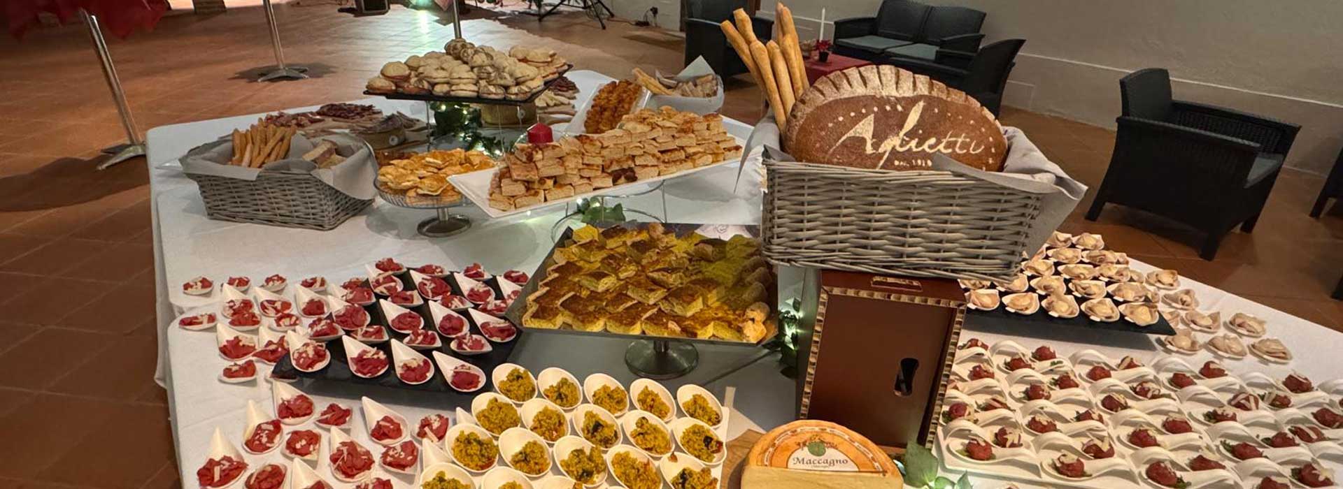 Just Grilled Catering by Aglietti presenta un ricco buffet di stuzzichini salati, allestito con cura su un tavolo rettangolare coperto da una tovaglia bianca. L'immagine mostra una varietà di finger food salati, panini e stuzzichini al cucchiaio, disposti in modo invitante su vassoi e alzatine di diverse forme e materiali. Un cesto di vimini contiene pane artigianale con il logo Aglietti dal 1910. Al di sotto di esso, si nota una forma di formaggio Maccagno. Sullo sfondo, si intravedono poltrone scure.  Delizia i tuoi ospiti con un buffet indimenticabile firmato Just Grilled Catering by Aglietti. Scoprire i nostri menu personalizzati e richiedere un preventivo per il tuo evento aziendale