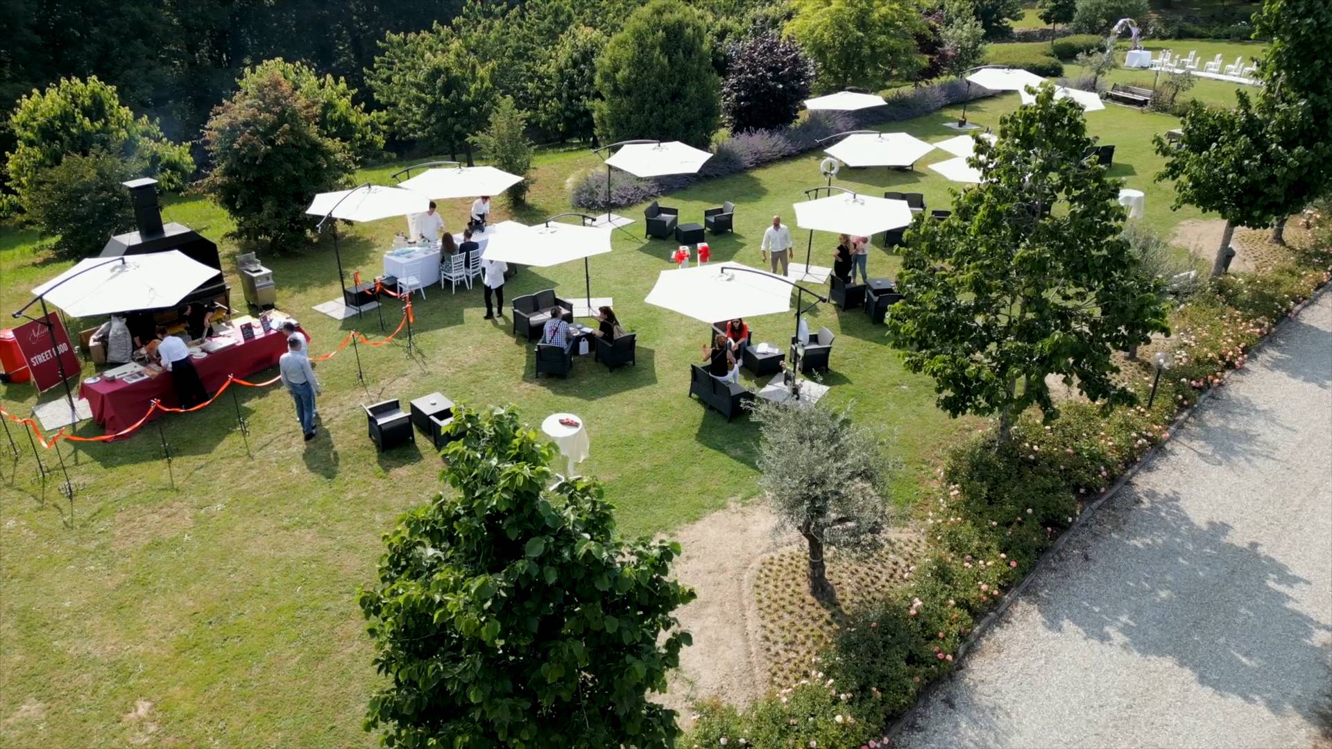 Just Grilled Catering by Aglietti presenta un evento all'aperto in un'ampia area verde. La vista aerea mostra un prato ben curato con alberi rigogliosi e aiuole fiorite. L'allestimento include numerosi ombrelloni bianchi che offrono ombra a tavolini con sedute scure. Un'area catering è chiaramente identificabile con un bancone rosso e personale di servizio, affiancata da un grande barbecue a legna. Ospiti che socializzano e si godono la giornata. Sullo sfondo, si distingue una suggestiva navata nuziale, completa di arco decorato e tavolo per il cerimoniere, pronta per celebrare un matrimonio da favola, tutto rigorosamente in binaco. Un sentiero in ghiaia conduce verso un'area più appartata del giardino. Organizza il tuo evento all'aperto perfetto con Just Grilled Catering by Aglietti. Scopri le nostre opzioni di menu e richiedere un preventivo personalizzato.